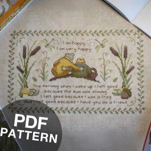 May include: Cross-stitch pattern featuring two frogs, one in a yellow coat, the other in green, with text that reads "I am happy, I am very happy." The design includes cattails and a decorative border. The words "PDF PATTERN" are also visible.
