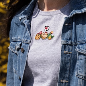 Op de afbeelding: Een grijs T-shirt met een gepixeld geborduurd ontwerp van twee cartoonfiguren die bloemen vasthouden en een spraakballon met een hart. Het shirt wordt gedragen door iemand die een spijkerjack draagt.
