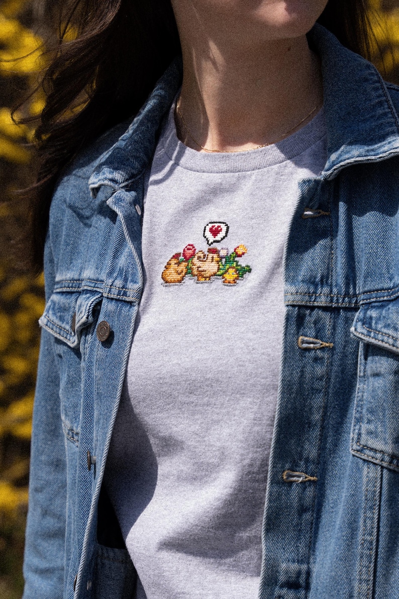 Op de afbeelding: Een grijs T-shirt met een gepixeld geborduurd ontwerp van twee cartoonfiguren die bloemen vasthouden en een spraakballon met een hart. Het shirt wordt gedragen door iemand die een spijkerjack draagt.