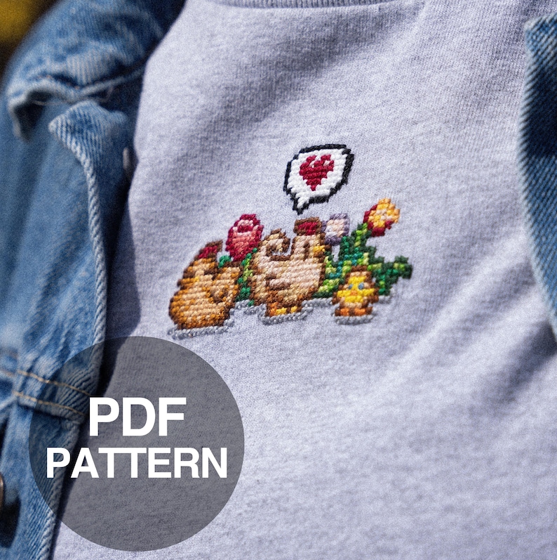 Op de afbeelding: Een grijs sweatshirt met een gepixelde kruissteekontwerp van twee cartoonfiguren die bloemen en een hart vasthouden. De tekst "PDF PATTERN" is afgedrukt op een cirkel in de rechterbenedenhoek van de afbeelding.