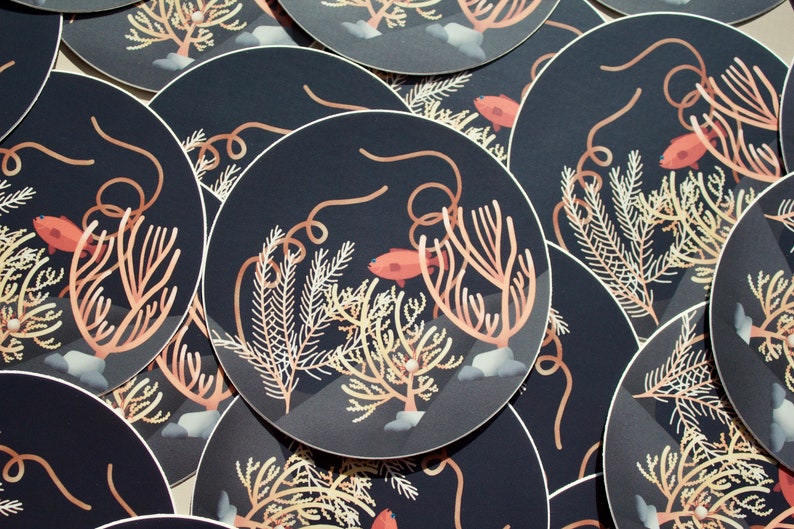 Op de afbeelding: Een set ronde stickers met een donkerblauwe achtergrond waarop koralen, zeewier en kleine oranje vissen zijn afgebeeld. De stickers zijn perfect om een ​​toets van zeeleven aan je spullen toe te voegen.