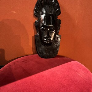 Puede incluir: Una m&aacute;scara de madera negra con un rostro humano y un tocado decorativo. La m&aacute;scara est&aacute; sentada sobre una superficie de terciopelo rojo.