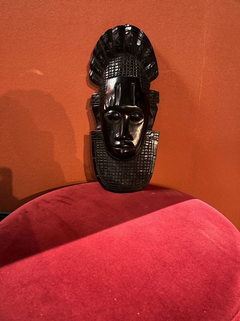 Puede incluir: Una m&aacute;scara de madera negra con un rostro humano y un tocado decorativo. La m&aacute;scara est&aacute; sentada sobre una superficie de terciopelo rojo.