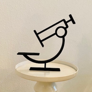 Op de afbeelding: Een zwart metalen beeldje van een microscoop op een witte sokkeltafel.