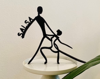 May include: Black metal silhouette of a couple dancing salsa on a white pedestal. The word "SALSA" is written in the silhouette.