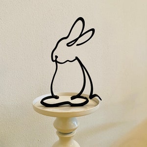 May include: A black metal rabbit sculpture on a white pedestal. The rabbit is in a sitting position with its ears up.