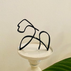 Könnte beinhalten: Eine schwarze Metallskulptur eines stilisierten Pferdes, das auf einem weißen Sockel steht.