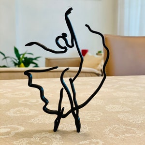 May include: A black metal sculpture of a ballerina in a graceful pose. The sculpture is made of thin wire and has a delicate, minimalist design.