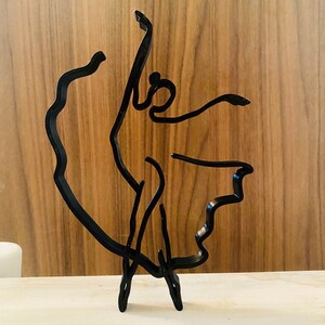 May include: Black metal sculpture of a dancer in a flowing pose. The sculpture is made of thin metal and has a minimalist design.