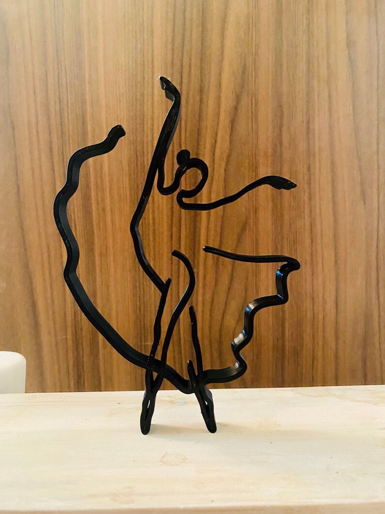 May include: Black metal sculpture of a dancer in a flowing pose. The sculpture is made of thin metal and has a minimalist design.