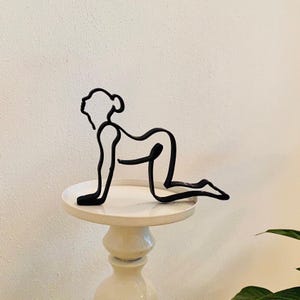 Peut inclure: Sculpture en métal noir d'une personne en position de yoga, sur un piédestal blanc.