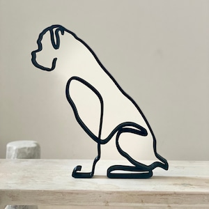 Könnte beinhalten: Schwarze Metalldrahtskulptur eines sitzenden Boxers mit nach unten geneigtem Kopf. Die Skulptur steht auf einer weißen Holzoberfläche.