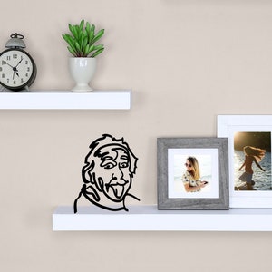 Puede incluir: Dos estantes blancos con un reloj despertador negro, una peque&ntilde;a planta en maceta, un dibujo en blanco y negro de un hombre con la lengua fuera y dos fotos enmarcadas en el estante superior.