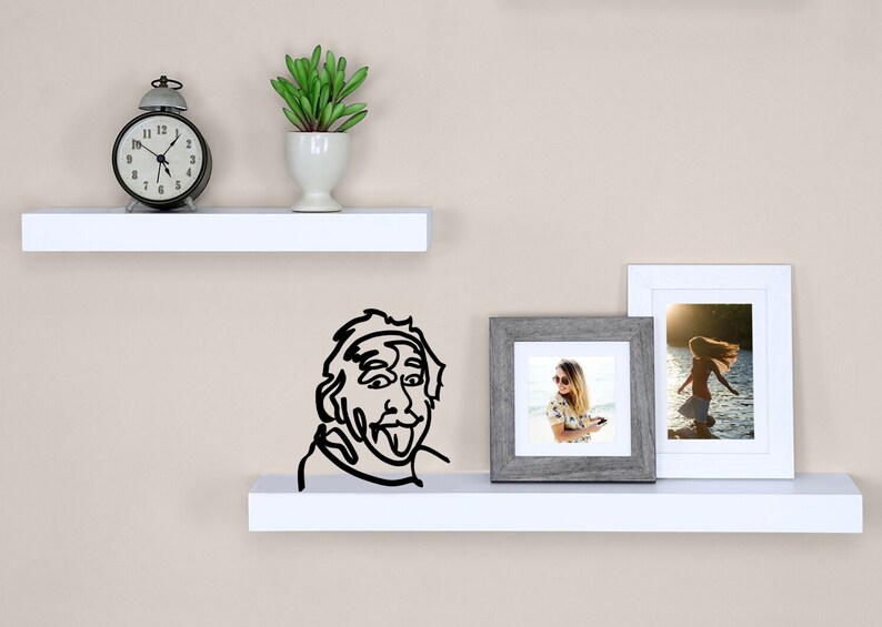 Puede incluir: Dos estantes blancos con un reloj despertador negro, una peque&ntilde;a planta en maceta, un dibujo en blanco y negro de un hombre con la lengua fuera y dos fotos enmarcadas en el estante superior.