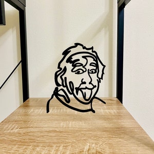 Puede incluir: Escultura de pared de metal negro de un rostro con la lengua fuera. La escultura est&aacute; en un estante de madera.