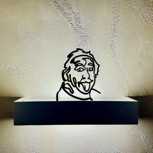 Puede incluir: Un dibujo en blanco y negro de una persona con la lengua fuera, montado en una estanter&iacute;a negra con una luz blanca detr&aacute;s.