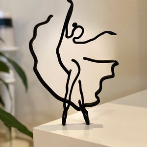 May include: Black metal sculpture of a dancer in a flowing dress. The sculpture is minimalist and abstract, with a simple line drawing style.