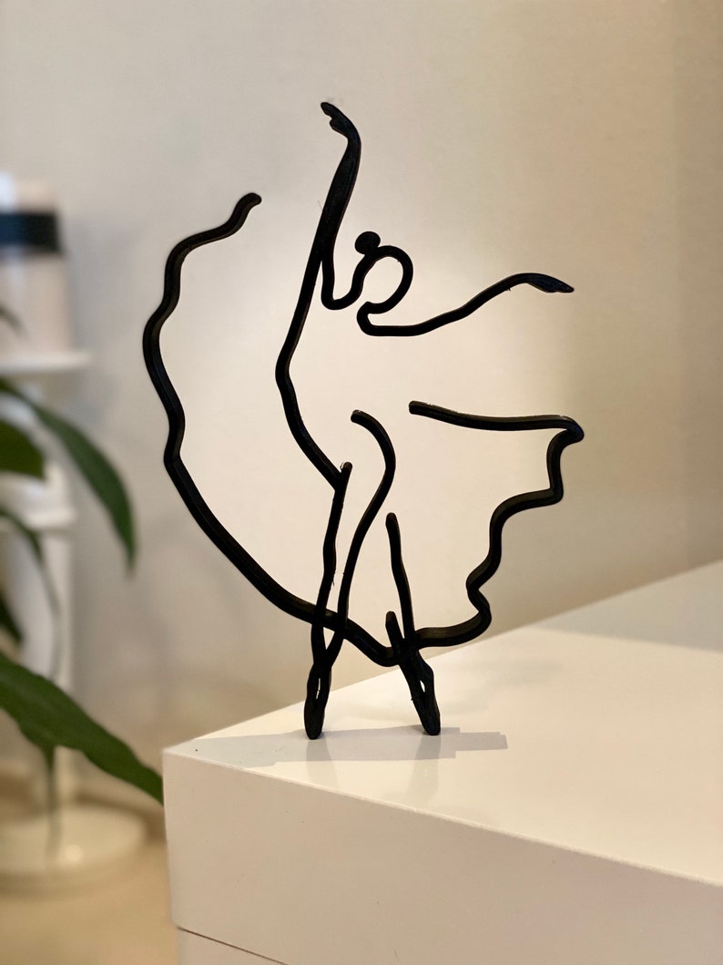 May include: Black metal sculpture of a dancer in a flowing dress. The sculpture is minimalist and abstract, with a simple line drawing style.