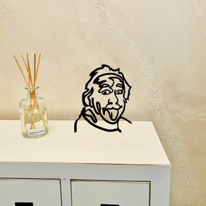 Puede incluir: Decoraci&oacute;n de pared de arte de l&iacute;nea en blanco y negro de una persona con la lengua fuera. El arte est&aacute; montado en un estante blanco con una botella de vidrio transparente y palos en el lado izquierdo.