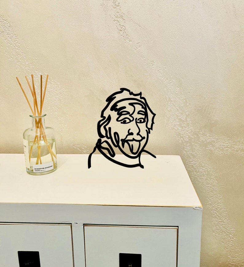 Puede incluir: Decoraci&oacute;n de pared de arte de l&iacute;nea en blanco y negro de una persona con la lengua fuera. El arte est&aacute; montado en un estante blanco con una botella de vidrio transparente y palos en el lado izquierdo.