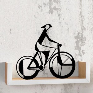 Peut inclure: Décoration murale en métal noir représentant un cycliste sur un vélo. Le design minimaliste montre une personne à vélo avec un chapeau. L'œuvre est montée sur une étagère blanche contre un mur texturé.