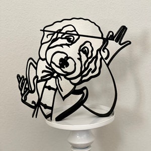 Puede incluir: Escultura de metal negro de un personaje de dibujos animados con pajarita, gafas y una cuerda. La escultura está montada sobre un pedestal blanco.