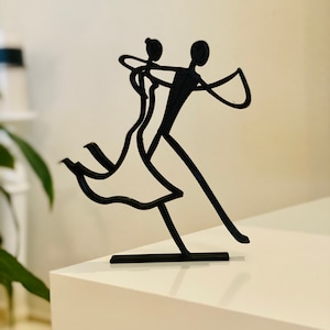 May include: Black metal sculpture of a couple dancing. The woman is wearing a dress and the man is holding her hand. The sculpture is on a white surface.