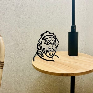 Puede incluir: Una l&aacute;mpara de mesa de metal negro con una mesa redonda de madera. Un dibujo de l&iacute;nea negra de una persona con la lengua fuera est&aacute; en la mesa.
