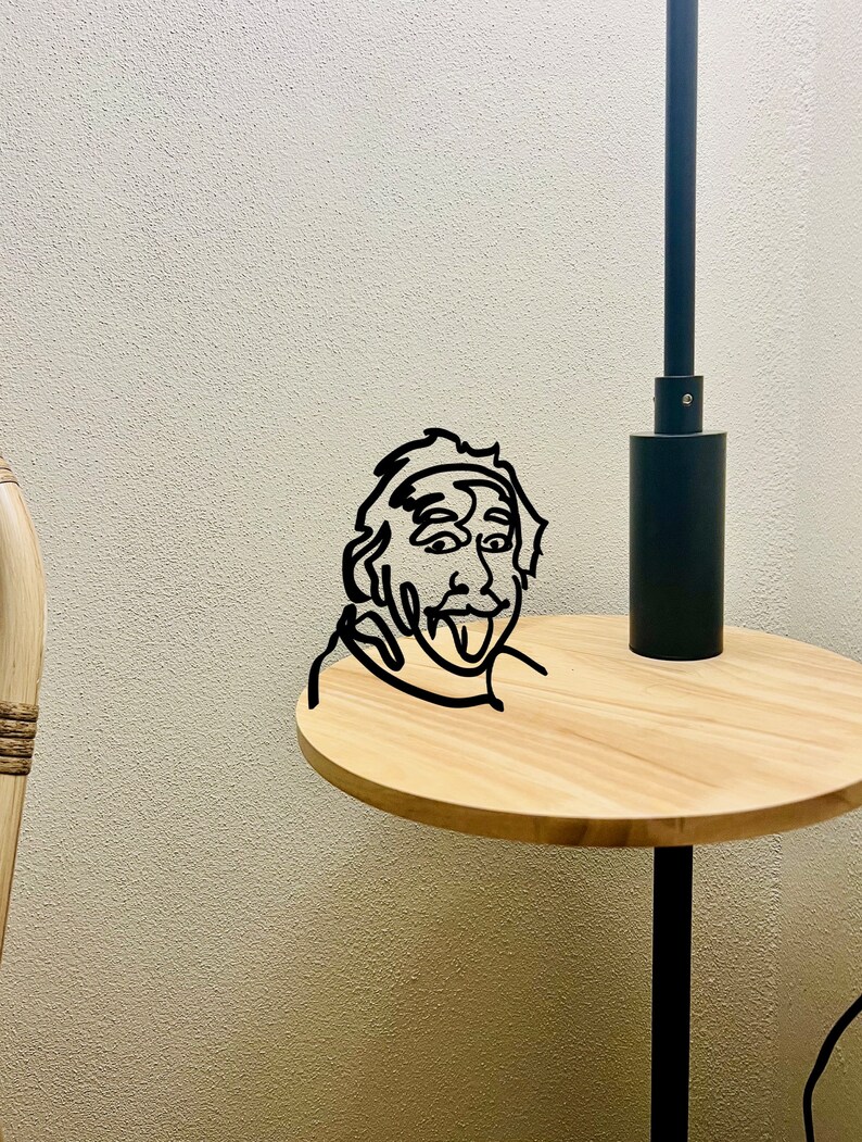 Puede incluir: Una l&aacute;mpara de mesa de metal negro con una mesa redonda de madera. Un dibujo de l&iacute;nea negra de una persona con la lengua fuera est&aacute; en la mesa.