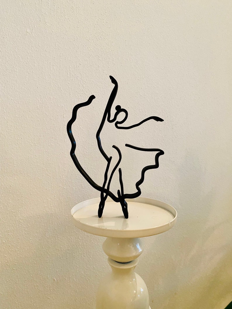 May include: Black metal sculpture of a ballerina in a dancing pose on a white pedestal.