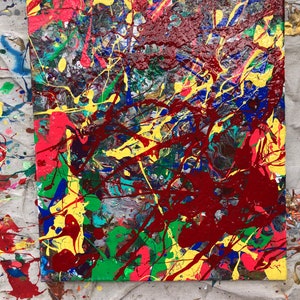 May include: Abstract painting on canvas with a colorful splatter design. The colors include red, yellow, blue, green, and black. The paint is splattered in a random pattern, creating a chaotic and energetic effect.