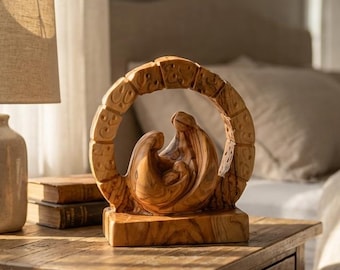May include: A carved olive wood sculpture depicting a stylized family unit enclosed within a circular frame with textured detailing, resting on a rectangular base. The wood features a natural grain pattern with light and dark brown tones.