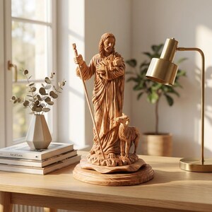 Op de afbeelding: Houten beeld van een religieuze figuur met een lam en een staf. De kleding en het gezicht van de figuur zijn gedetailleerd gesneden. Het beeld staat op een houten voet, op een houten tafel met een lamp en boeken.