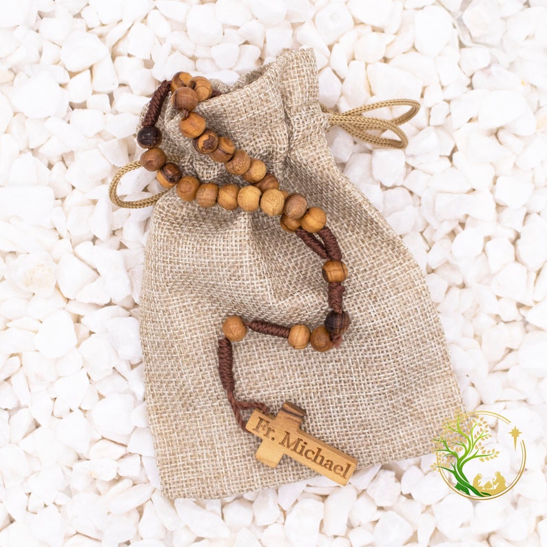 May include: A brown wooden rosary with a wooden cross engraved with "Fr. Michael" in a burlap drawstring bag. The bag is on a white background.