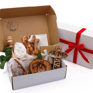 May include: A gift box with religious items, including a wooden cross, a carved nativity scene, and a rosary. The box also contains a white angel figurine and a wooden candle holder. A separate white box with a red ribbon is also present.