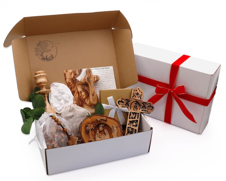 May include: A gift box with religious items, including a wooden cross, a carved nativity scene, and a rosary. The box also contains a white angel figurine and a wooden candle holder. A separate white box with a red ribbon is also present.
