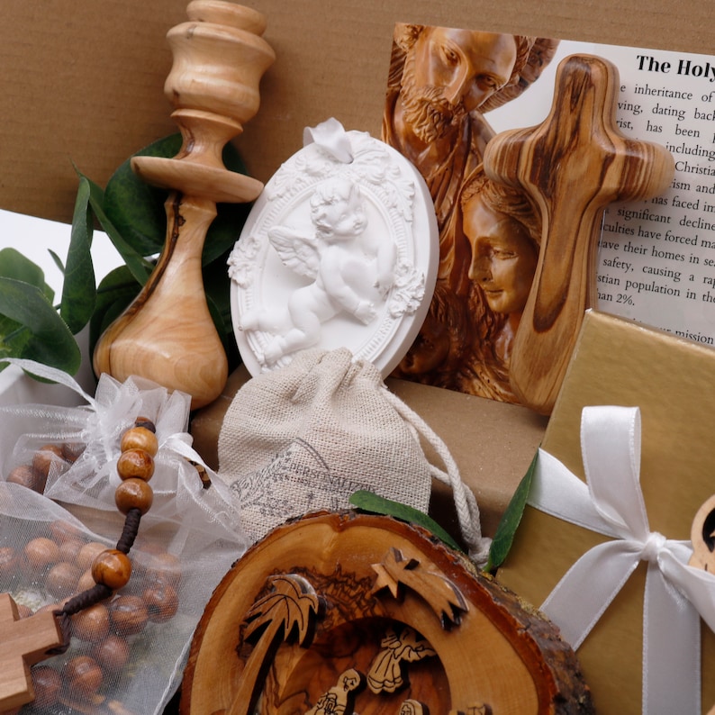 May include: A collection of religious items, including a wooden cross, a carved wooden nativity scene, a white angel relief, and a rosary. A wooden candlestick and a small gift box with a white ribbon are also present. The items are arranged in a box.