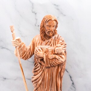 Op de afbeelding: Een gesneden houten beeld van een figuur met lang haar en een baard, die een staf en een lam vasthoudt. Het beeld heeft een lichtbruine kleur en staat tegen een witte marmeren achtergrond.