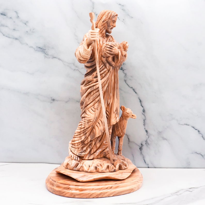 Op de afbeelding: Een gesneden houten beeld van een religieuze figuur die een lam vasthoudt, met een staf in de hand. De figuur draagt ​​losse gewaden en staat op een houten voet. Het beeld is lichtbruin.