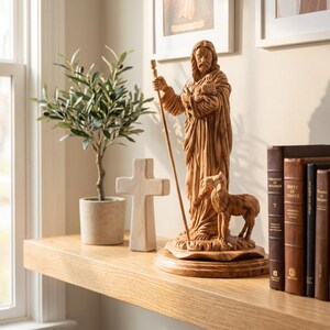 Op de afbeelding: Een houten beeld van Jezus met een lam en staf, met een klein lam aan zijn voeten. Een wit kruis, een plant in pot en boeken staan op een houten plank. Twee ingelijste foto's hangen aan de muur.