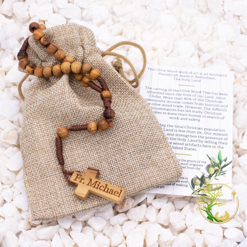 May include: A brown wooden rosary with a cross engraved with "Fr. Michael" is displayed in a burlap bag. The rosary is made from olive wood from the Holy Land. The text on the card reads: "This Olive Wood work of art is an Authentic Handicraft made in Bethlehem. The Holy Land."