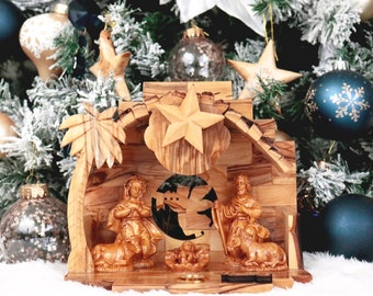 May include: A wooden nativity scene with a star on top, a manger with a baby Jesus, Mary and Joseph, and a sheep. The scene is set against a snowy Christmas tree background.