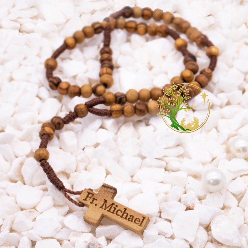 May include: A brown wooden rosary with a brown cord and a wooden cross with the inscription "Fr. Michael". The rosary is on a white background.