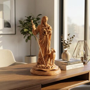 Op de afbeelding: Een lichtbruin houten beeld van een religieuze figuur die een lam en staf vasthoudt, met een schaap aan zijn voeten. Het beeld staat op een ronde basis en staat op een houten bureau.