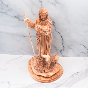Op de afbeelding: Een houten beeld van een religieuze figuur met een staf en een lam. De figuur, met lang haar en een baard, draagt een gewaad. Het beeld staat op een houten voet, met een lam aan zijn voeten.