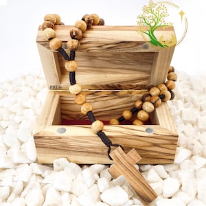 May include: A wooden box with a hinged lid, open to reveal a wooden rosary with brown beads and a wooden cross. The box is made of olive wood and has a dark brown stain. The box is sitting on a bed of white rocks.