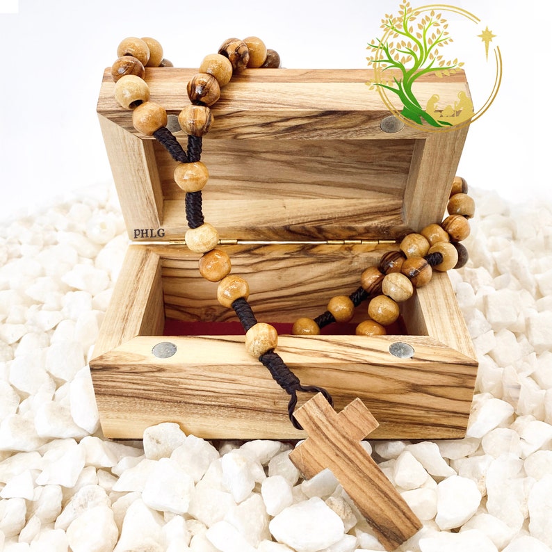 May include: A wooden box with a hinged lid, open to reveal a wooden rosary with brown beads and a wooden cross. The box is made of olive wood and has a dark brown stain. The box is sitting on a bed of white rocks.