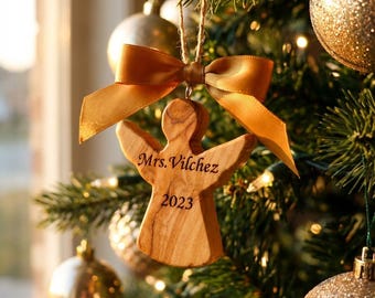 May include: A wooden angel Christmas ornament with the text "Mrs. Vilchez 2023" is suspended from a gold ribbon bow. The ornament is hanging on a Christmas tree with gold baubles and lights.