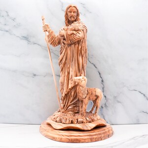 Op de afbeelding: Een houten beeldhouwwerk dat een figuur voorstelt die een staf en een lam vasthoudt, met een ander lam aan zijn voeten. Het beeld is gemaakt van lichtbruin hout en staat op een ronde, getextureerde basis. De figuur draagt een gewaad.