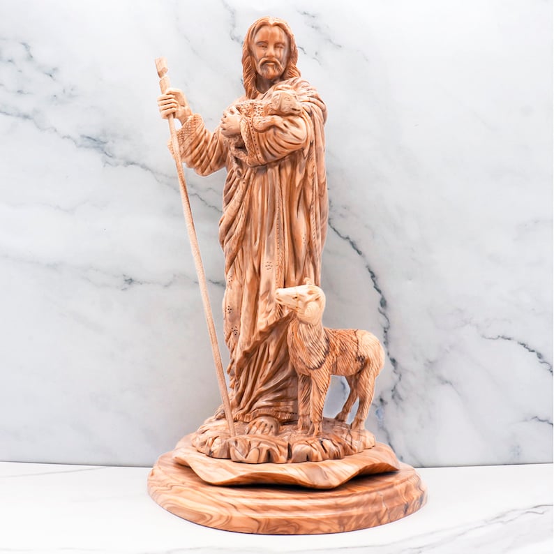 Op de afbeelding: Een houten beeldhouwwerk dat een figuur voorstelt die een staf en een lam vasthoudt, met een ander lam aan zijn voeten. Het beeld is gemaakt van lichtbruin hout en staat op een ronde, getextureerde basis. De figuur draagt een gewaad.